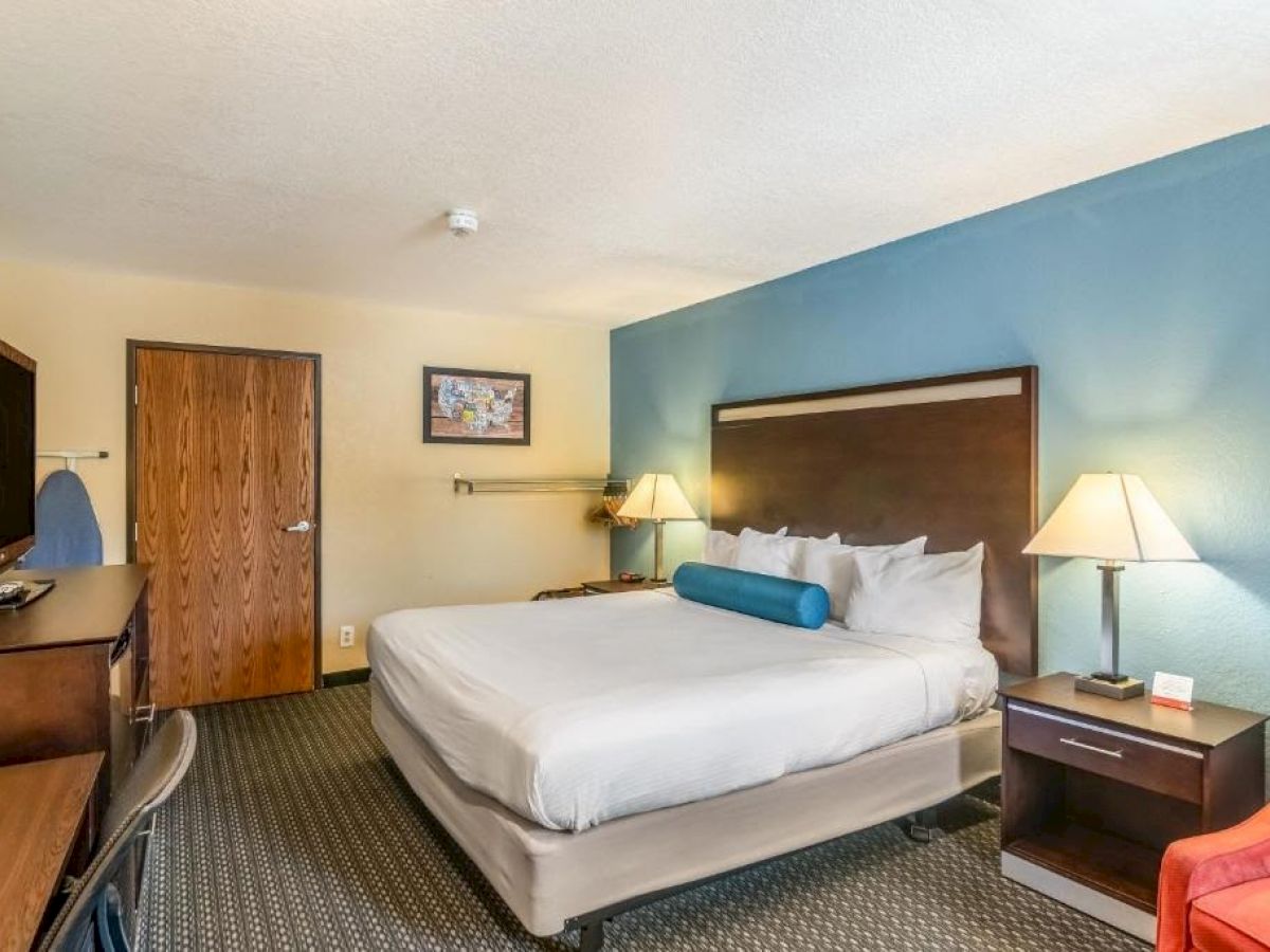 A hotel room with a large bed, wooden furniture, a blue accent wall, desk, chair, two lamps, and a framed picture near the door.
