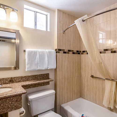 A modern bathroom features a granite countertop, a large mirror, a towel rack, a toilet, and a shower-tub combo with tiled walls and a curtain.