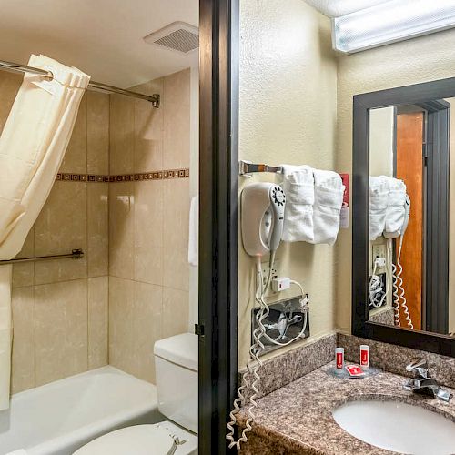 A bathroom with a tub-shower combo, beige tiles, a granite countertop with a sink, a mirror, a hairdryer, and a glimpse into a bedroom ends the sentence.
