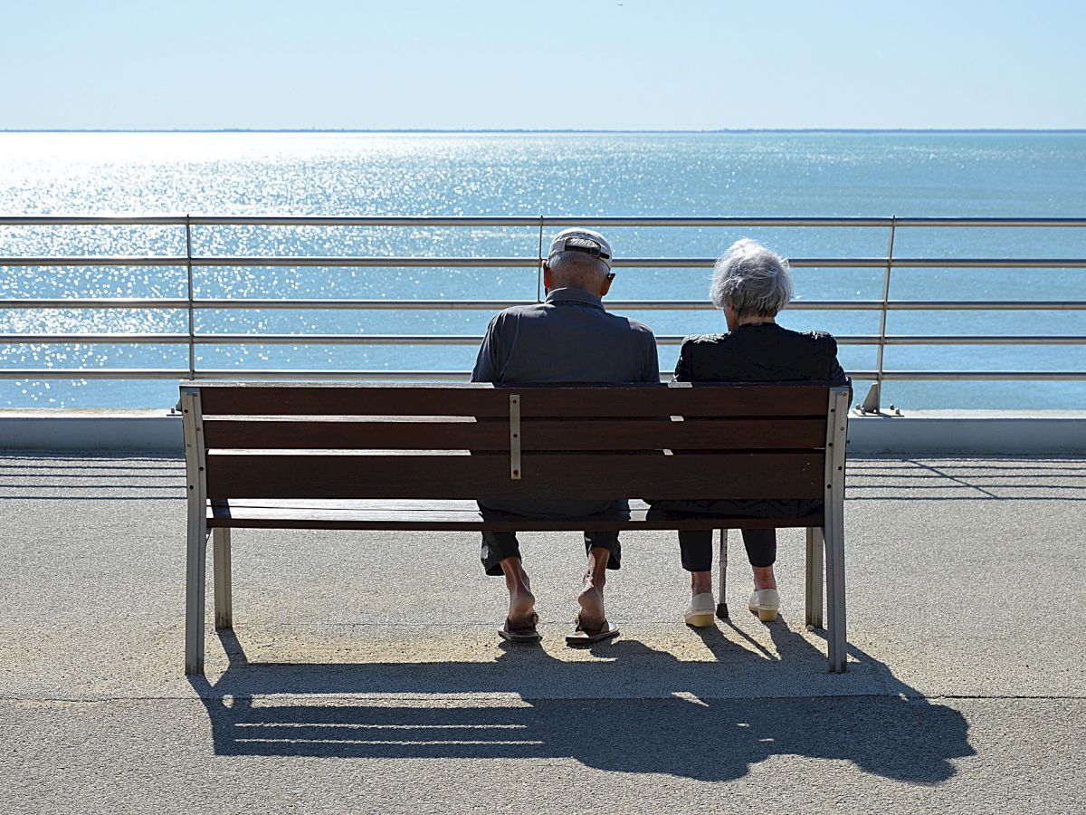 An elderly couple sits on a bench by the seaside, gazing at the calm ocean under a clear blue sky, enjoying each other's company.