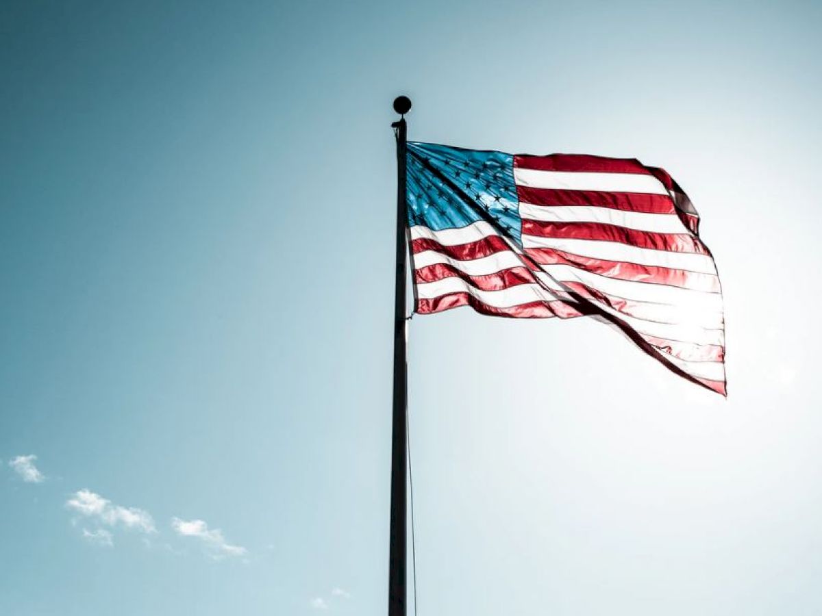 An American flag waves against a clear blue sky, illuminated by sunlight from behind, with slight clouds visible in the background.
