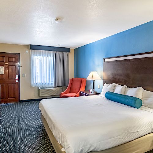 A hotel room with a large bed, blue accent wall, TV, dresser, red chair, table lamps, and a window with curtains.