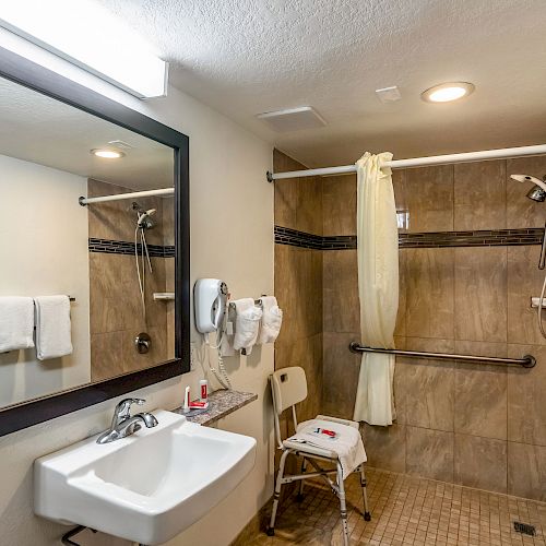 This image shows a bathroom equipped with accessibility features, including a shower chair, grab bars, and a sink with a large mirror.