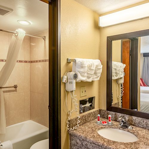 A hotel bathroom with a bathtub, sink, mirror, towels, and toiletries. A hairdryer is mounted on the wall, and a glimpse of the bedroom is visible.