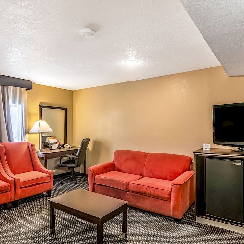 The image shows a hotel room with red seating, a desk with a chair, a table, a TV on a cabinet, and a microwave.