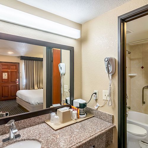 The image shows a bathroom with a granite countertop, toiletries, hairdryer, and a large mirror reflecting a bedroom. A shower curtain is partly shown.