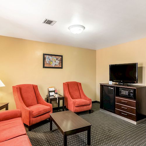 This image shows a cozy hotel room with red furniture, a TV, a lamp, and a table. There's a bedroom visible through an open door.