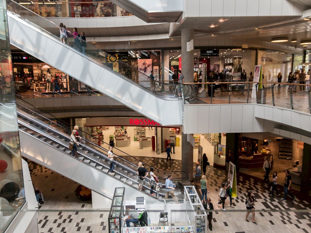 The image shows the interior of a multi-level shopping mall with escalators, various stores, and people shopping and walking around.