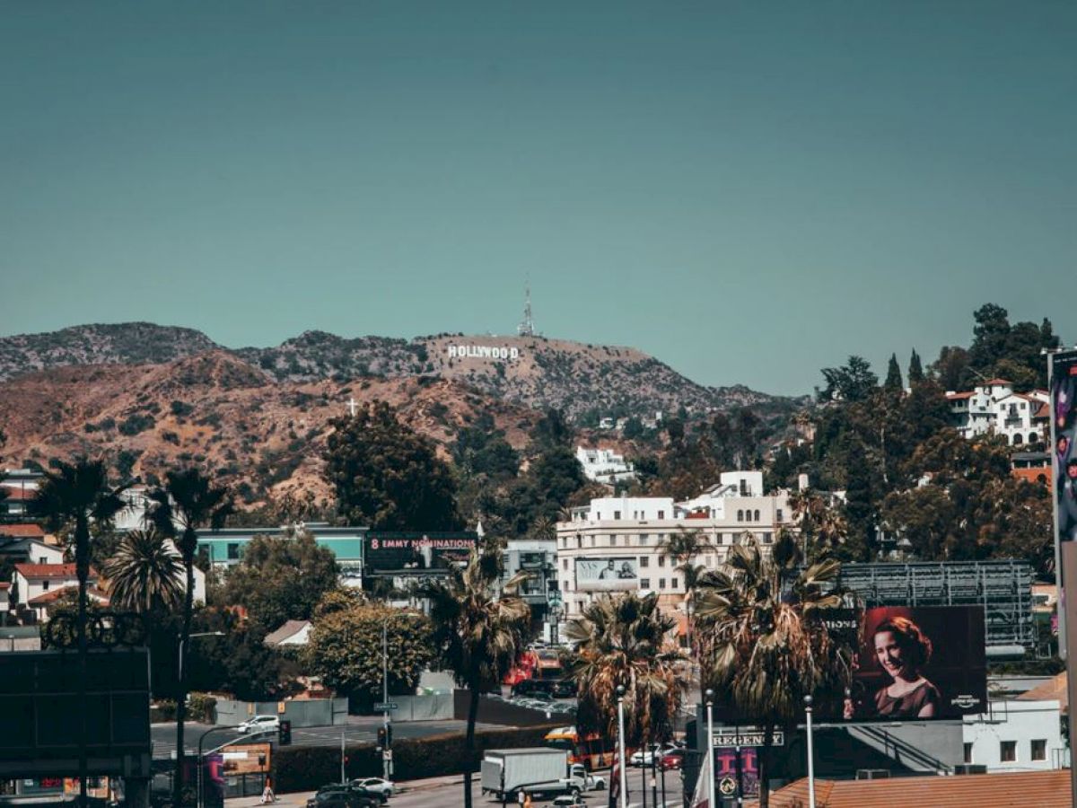 The image shows a cityscape with the iconic Hollywood sign in the background, palm trees, buildings, and billboards under a clear sky.
