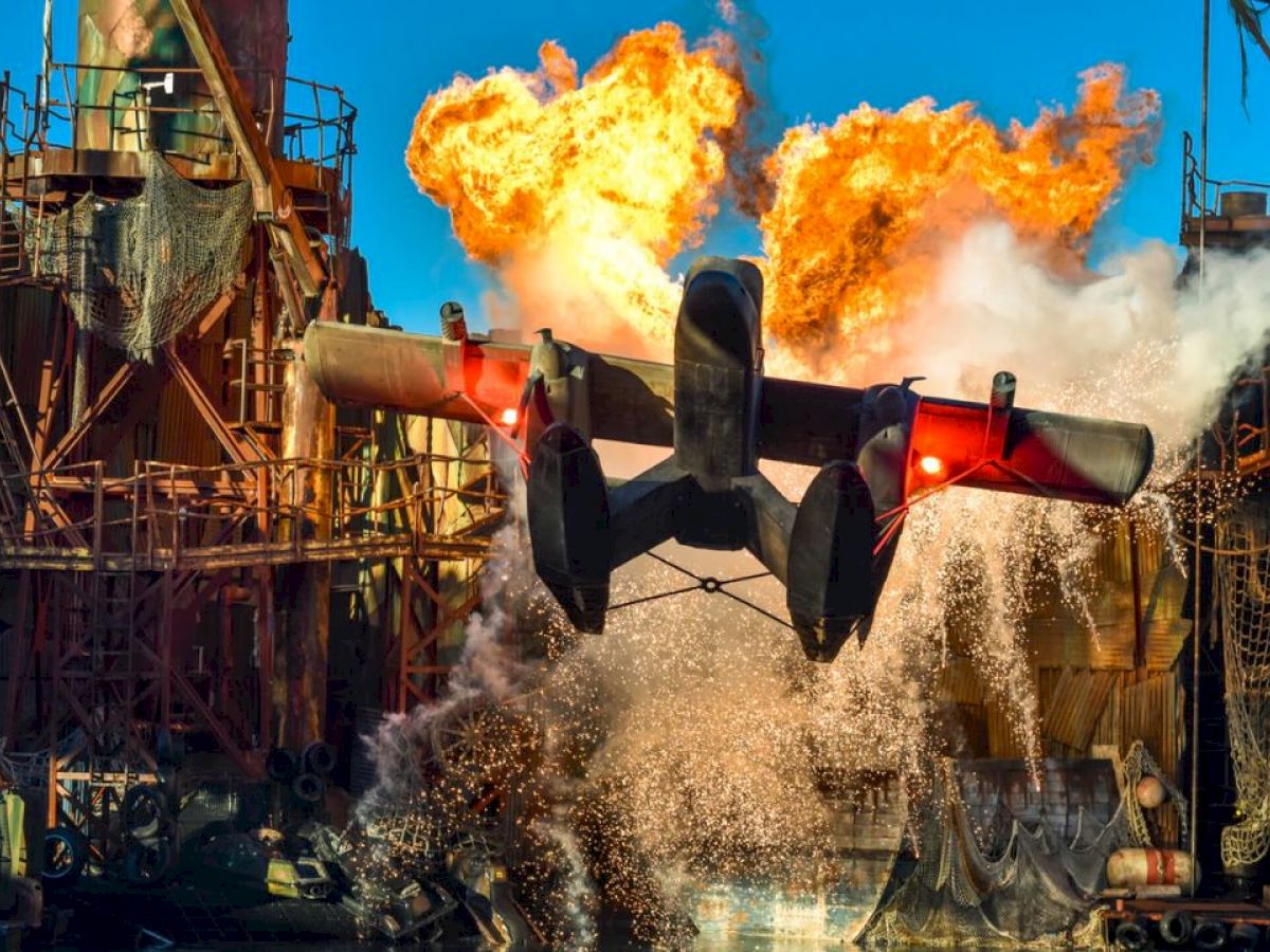 A seaplane is taking off from water amidst fiery explosions in an industrial setting, with structures and fiery blasts in the background.