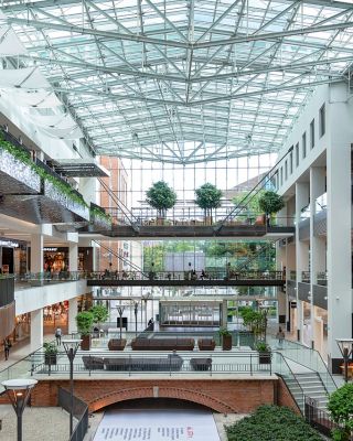 The image shows a spacious, multi-level shopping mall with a glass roof, various shops, green plants, and a modern design featuring open walkways.